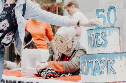 Passantin wirft eine Aktionspostkarte an Primark in eine Box in Form einer Geburtstagstorte. Im Hintergrund das Straßentheater vor dem Primark Münster