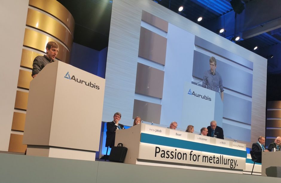 CIR-Referent Christian Wimberger steht auf dem Podium und hält eine Rede, links daneben sitzen die Aufsichtsräte und Vörstände von Aurubis, darüber eine große Leinwand, auf der der Referent nochmal zu sehen ist.