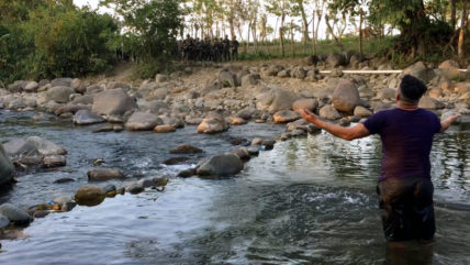 Aktivist steht im Fluss Guapinol und breitet die Arme aus richtung einer bewaffneten Truppe Sicherheitskräfte in Uniform am anderen Ufer.
