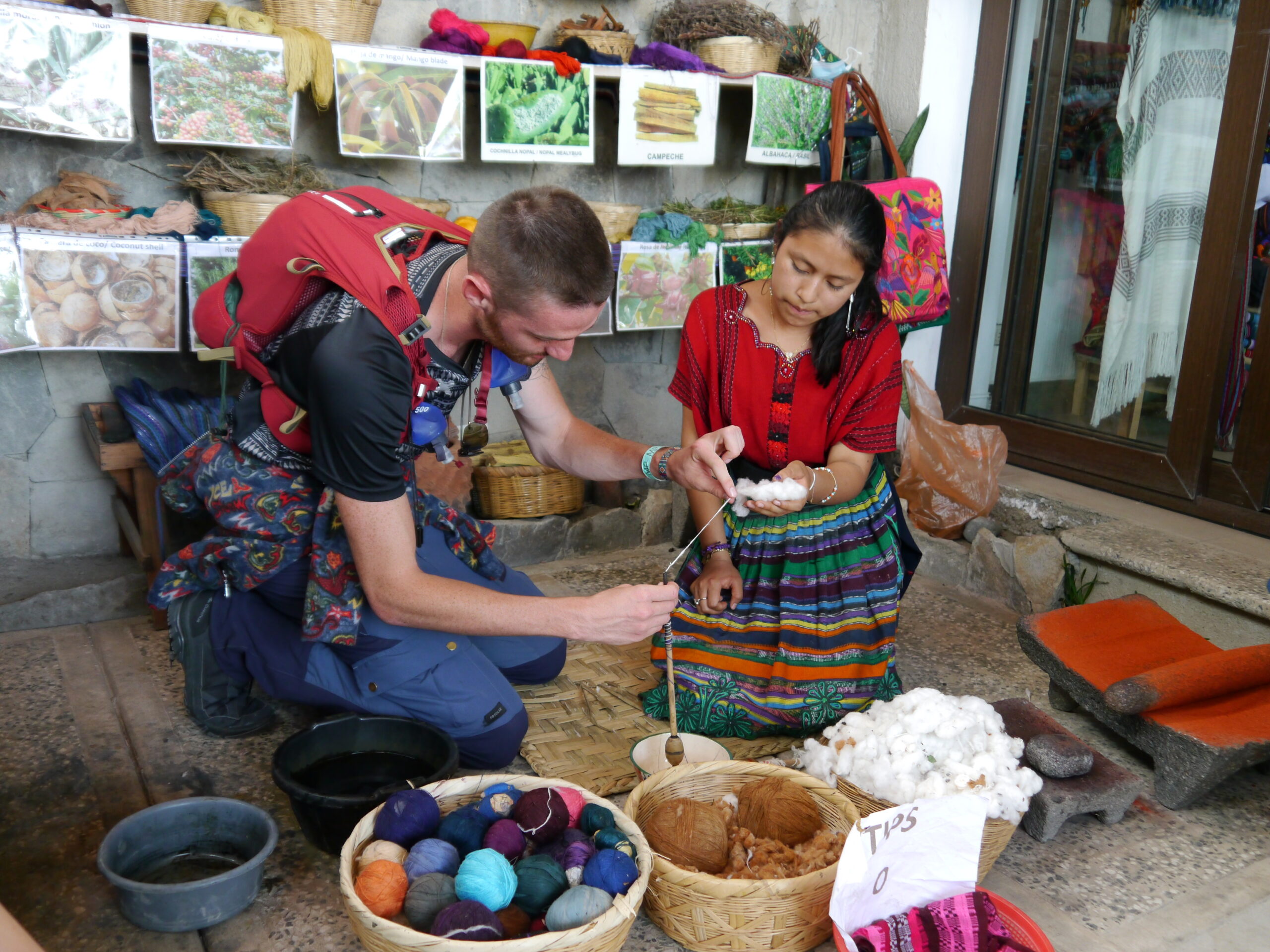 Eine Frau in traditioneller Maya-Kleidung und ein junger Mann knien auf dem Boden, vor ihnen Körbe mit bunten Wollknäueln und eine Spindel. Sie zeigt ihm, wie er den Baumwollfaden spinnen muss.