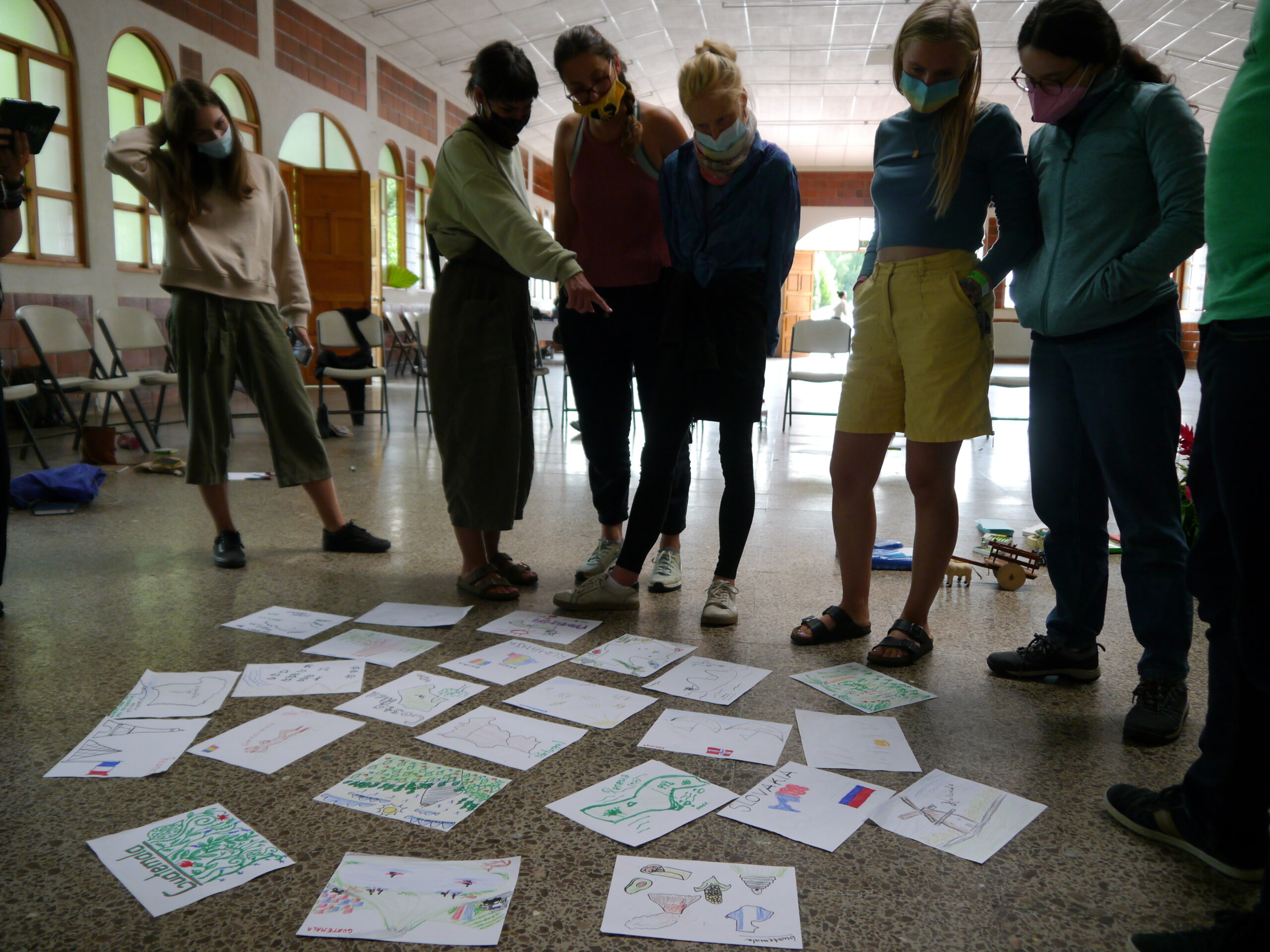 Sieben junge Menschen stehen in einem Workshopraum über auf dem Boden ausgebreiteten Bildern, eine junge Frau zeigt auf eins der Bilder