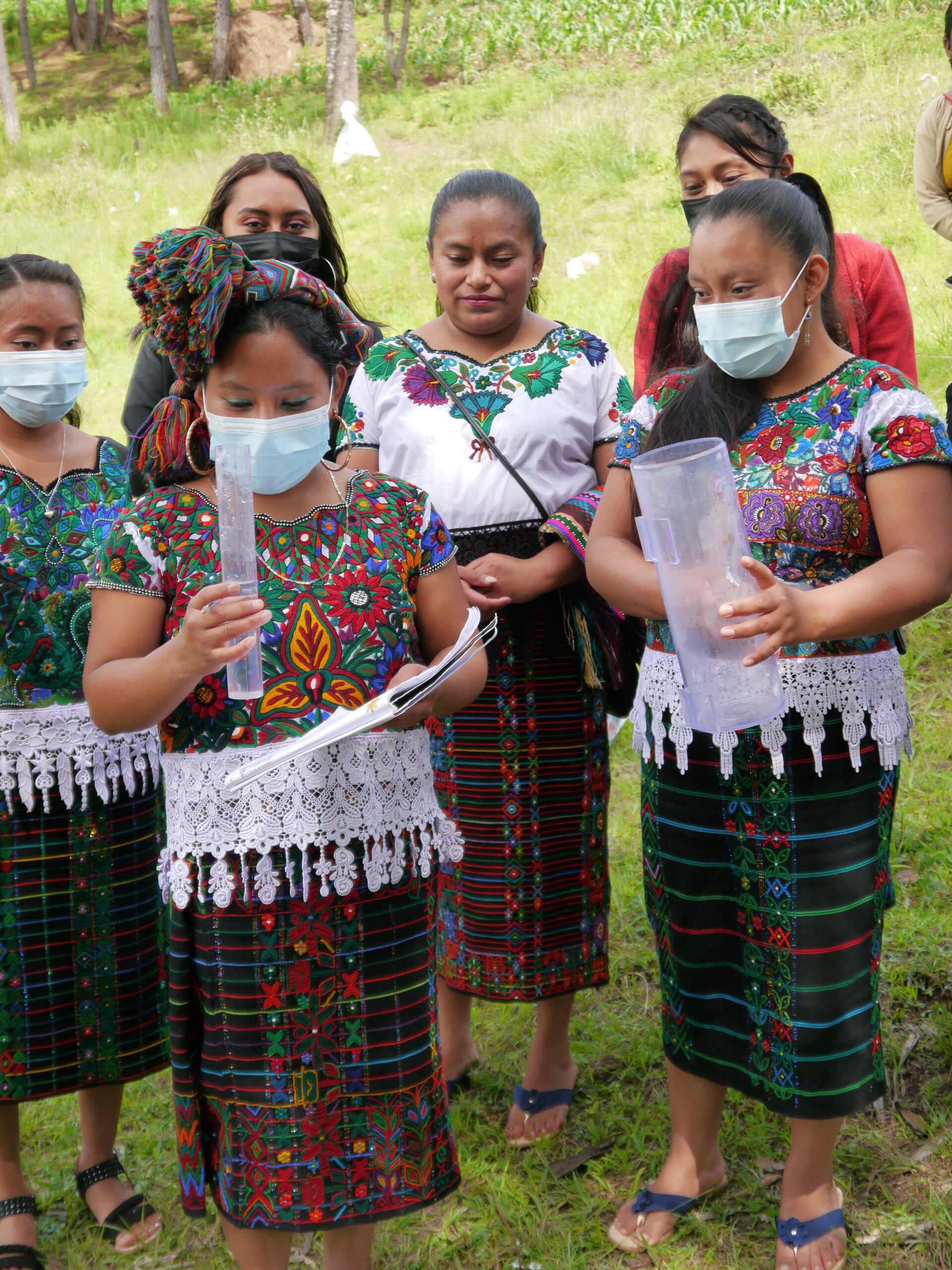 Eine Gruppe junger Frauen in traditioneller Maya-Kleidung steht um zwei Frauen herum. Sie halten Instrumente zur Messung von Niederschlagsmengen in den Händen
