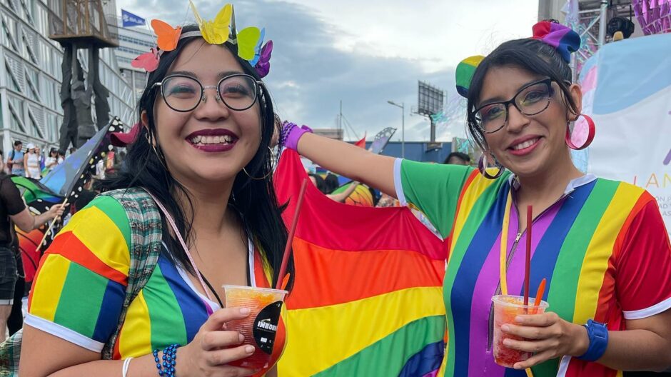 Zwei Personen sind in regenbogenfarben gekleidet und halten eine Regenbogenfahne. Beide tragen eine Brille und sind geschminkt.