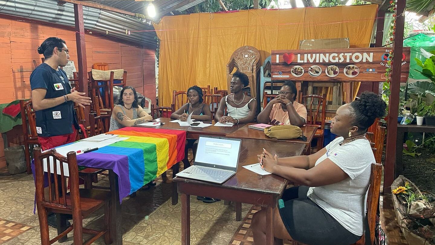 5 Menschen sitzen an mehreren Tischen und hören einer stehenden Person zu. Auf dem einen Tisch liegt eine Regenbogenflagge.