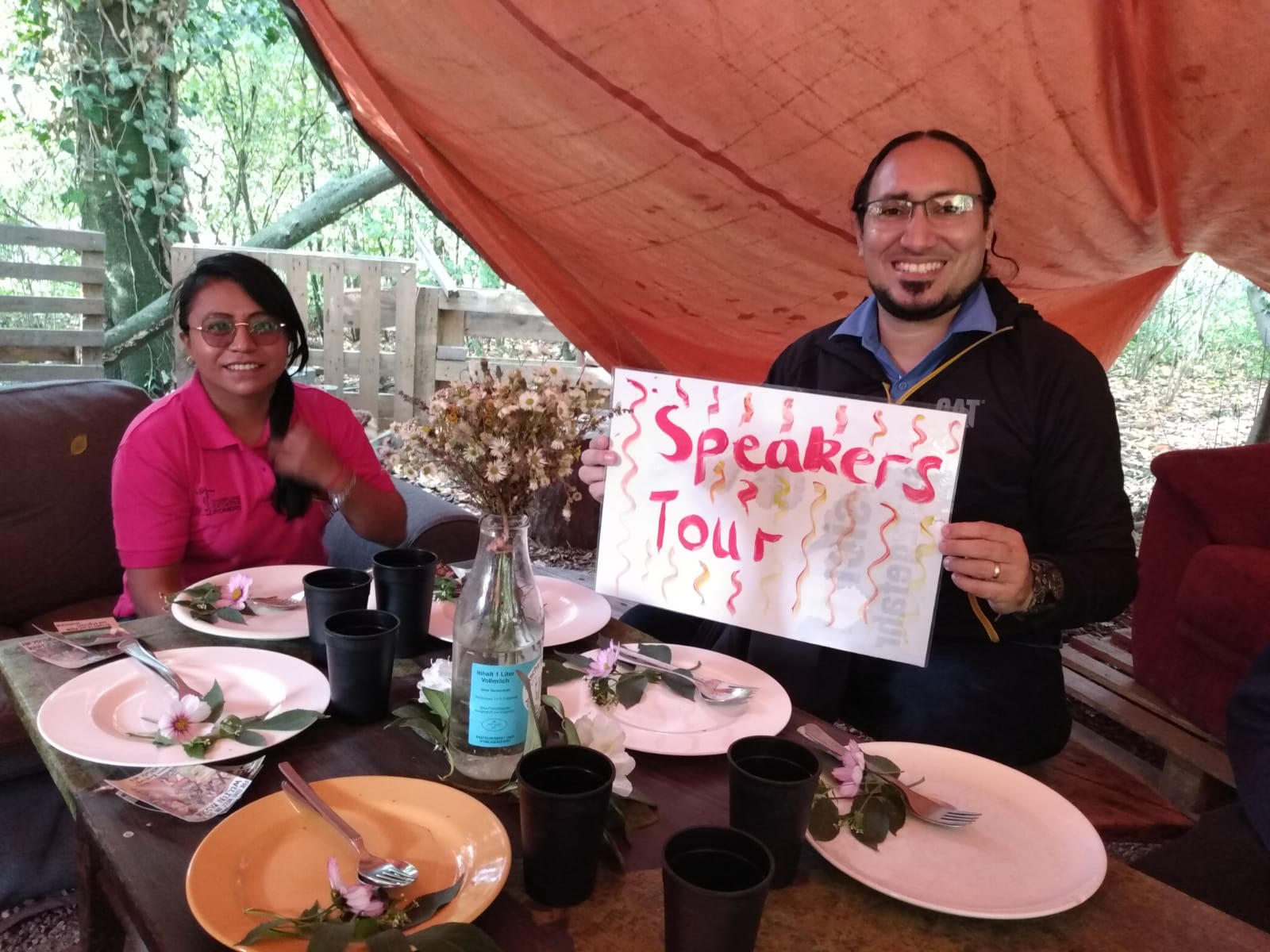 Luis González und Marlen Corea sitzen an einem gedeckten Esstisch und Luis González hält ein Schild mit den Worten "Speakers Tour". Beide lächeln,