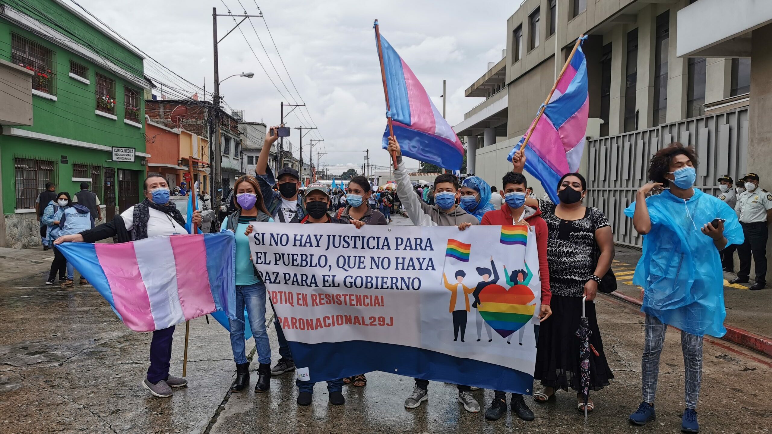 Demonstrierende tragen OP-Masken und halten ein Plakat auf dem "Si no hay justica para el pueblo, que no haya paz para el gobieno" steht. Außerdem werden drei blau-rosa-weiße Fahnen geschwenkt.