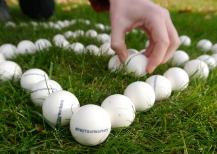 Auf dem Bild ist die Ecke eines Fußballfeldes zu sehen, welches auf Tischkicker-Bällen geformt wurde. Auf dem Bällen steht #PayYourWorkers. Eine Hand im Hintergrund legt einen weiteren Tischkicker-Ball auf das Feld.