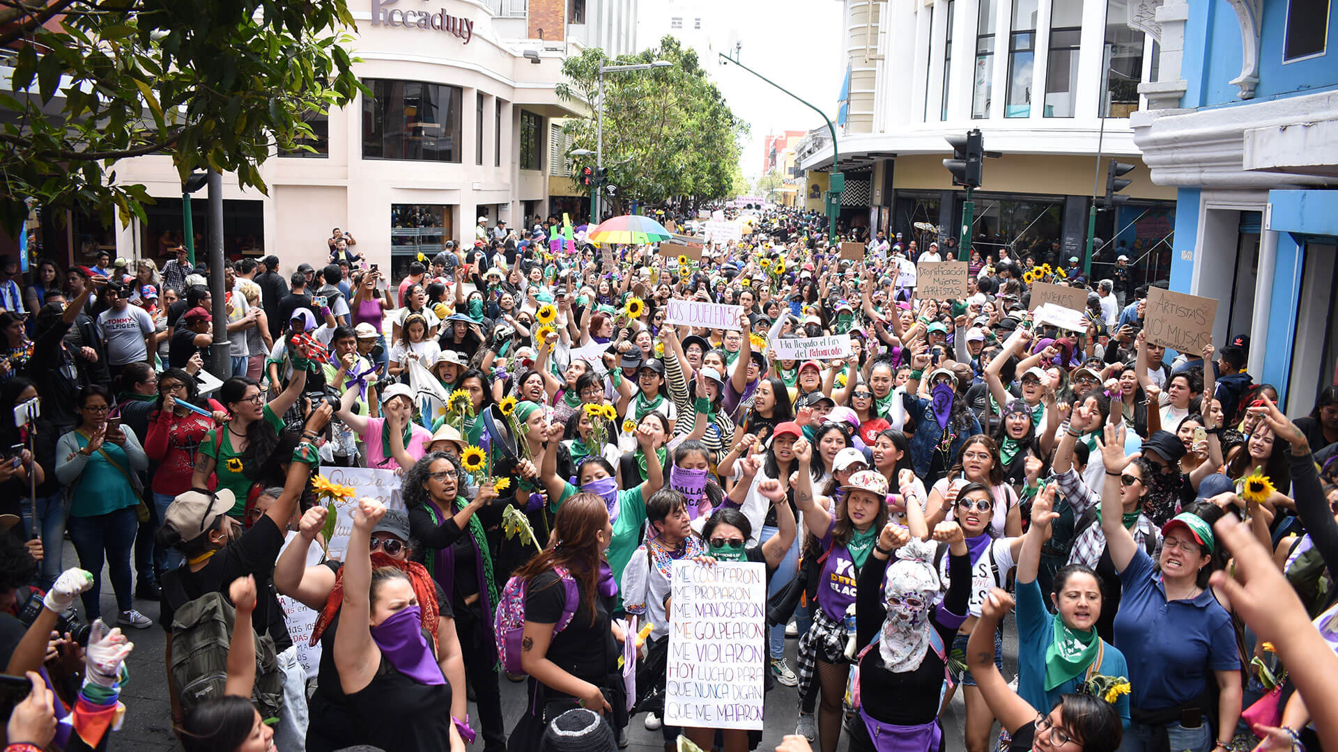 Demonstrierende mit hochgestreckten Fäusten, Schildern oder Sonnenblumen. Viele tragen die Farbe lila als Kleidung.