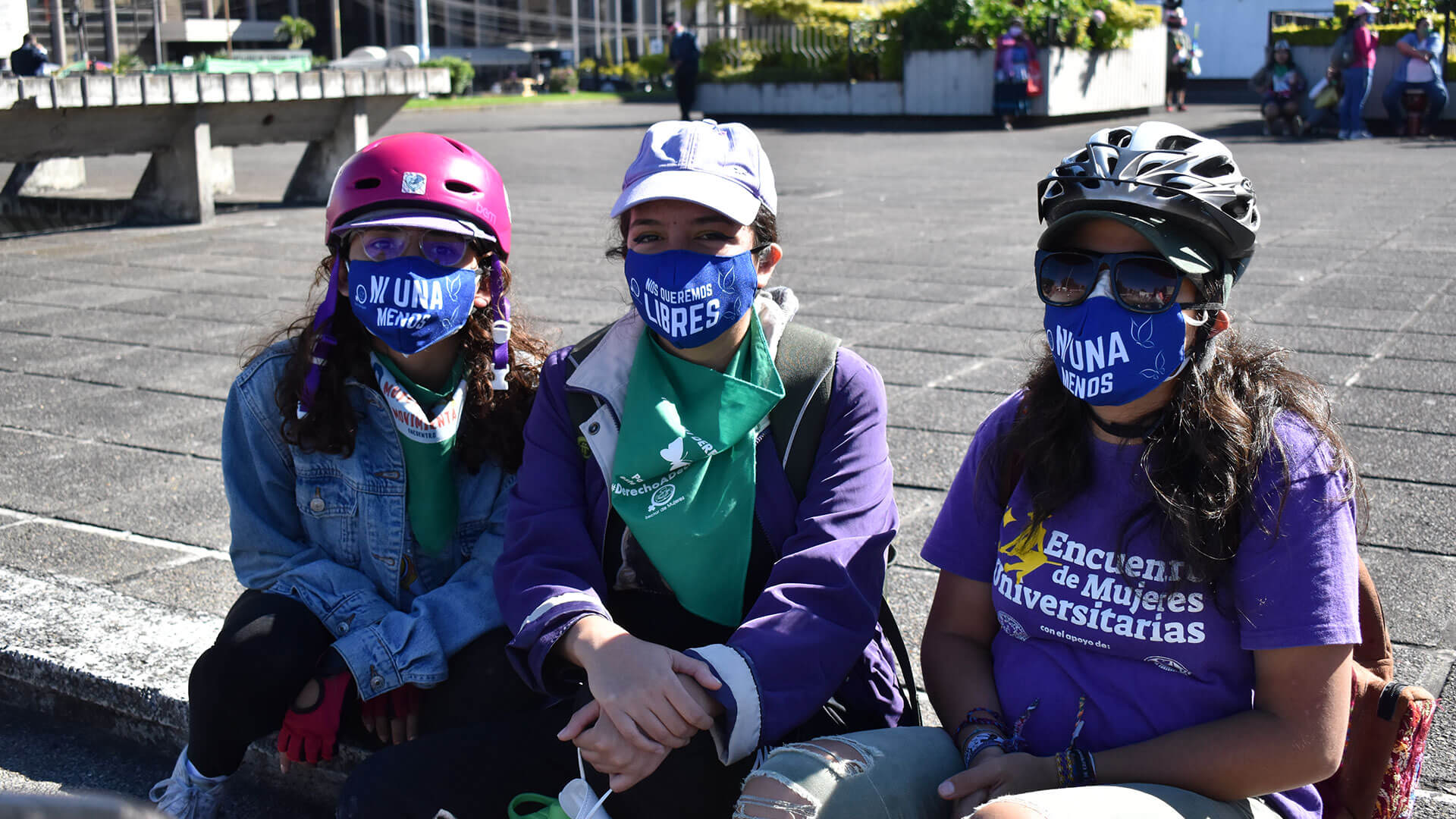 Drei Personen sitzen mit Masken "Ni una menos" auf einer Treppenstufe in der Öffentlichkeit.