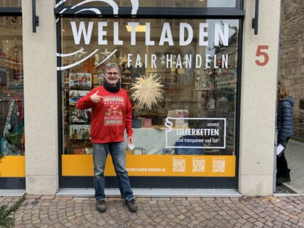 Herr Werum steht vor dem Schaufenster seines Weltladens und zeigt mit dem Daumen nach oben.