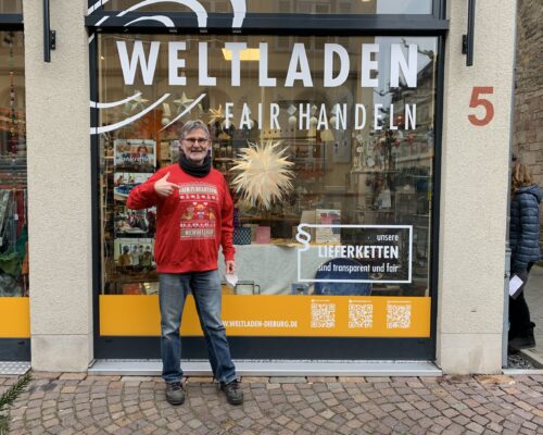 Herr Werum steht vor dem Schaufenster seines Weltladens und zeigt mit dem Daumen nach oben.
